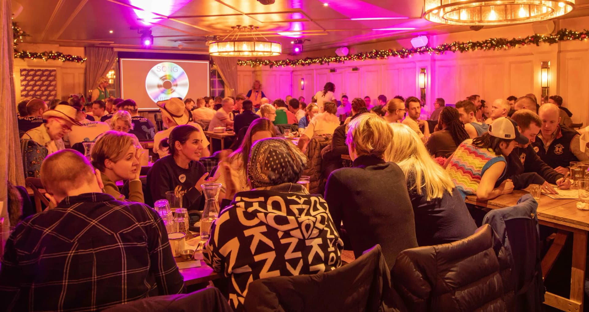 A packed, pink-lit room of teams mid-quiz.