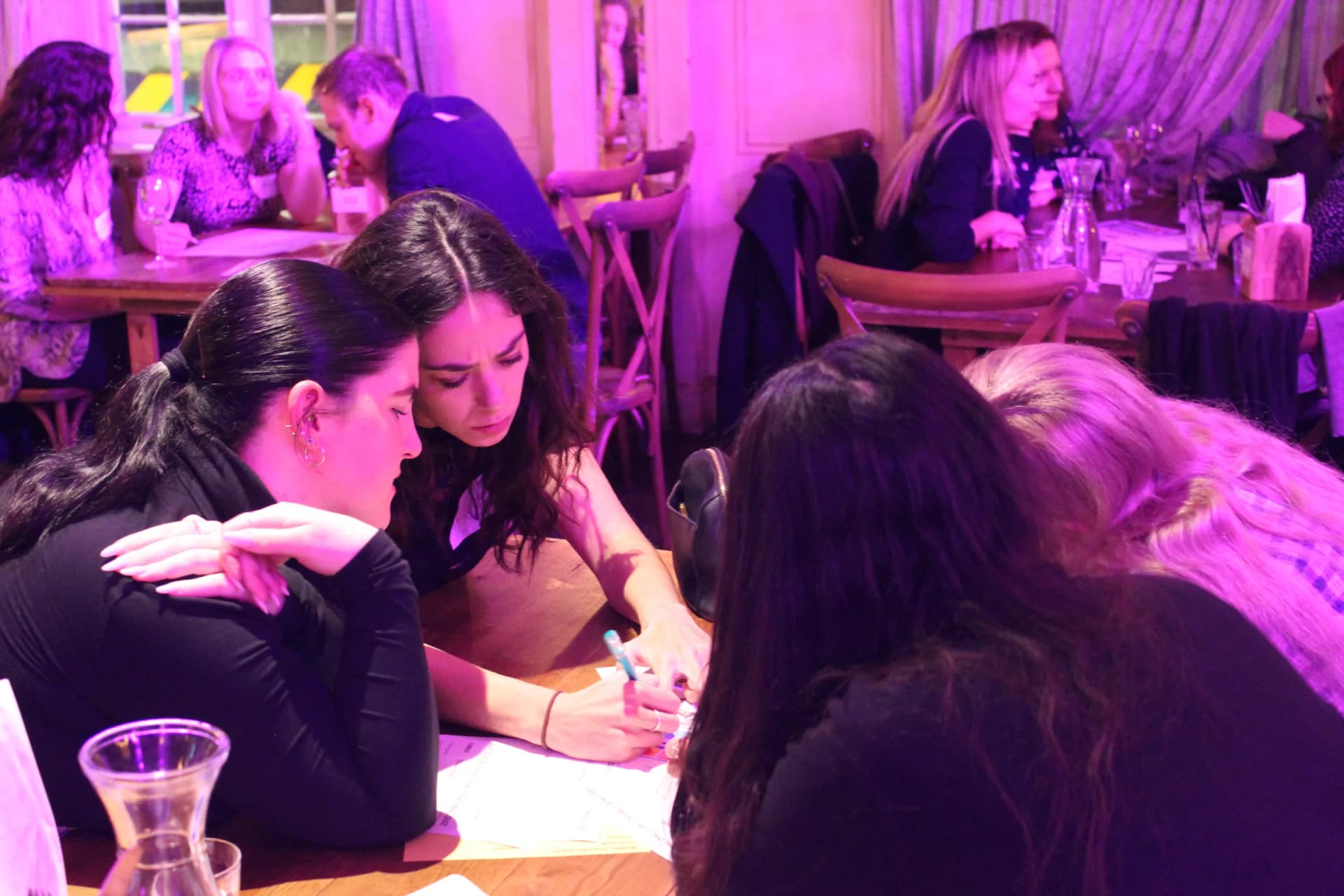 Two teammates heads together, writing an answer under pink light.