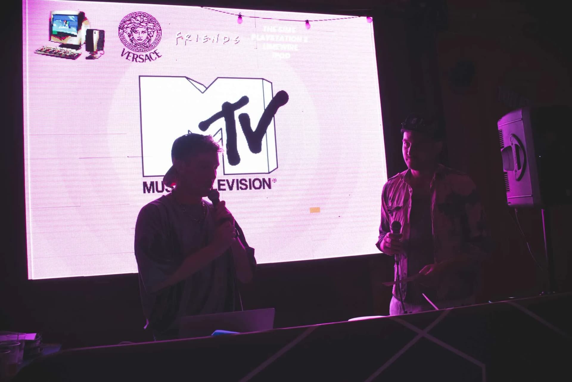 Hosts silhouetted against a retro music-TV logo on screen.