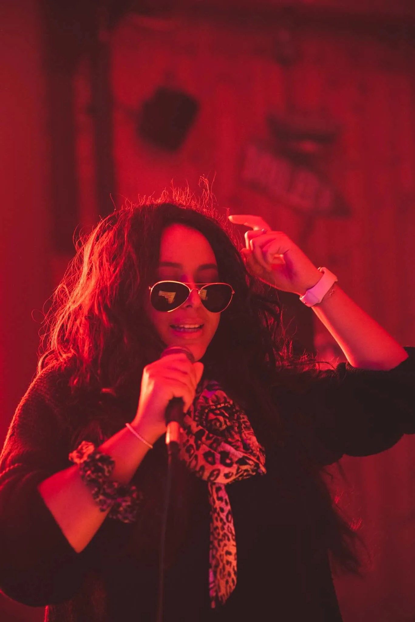 Singer in heart sunglasses mid-song under red light.