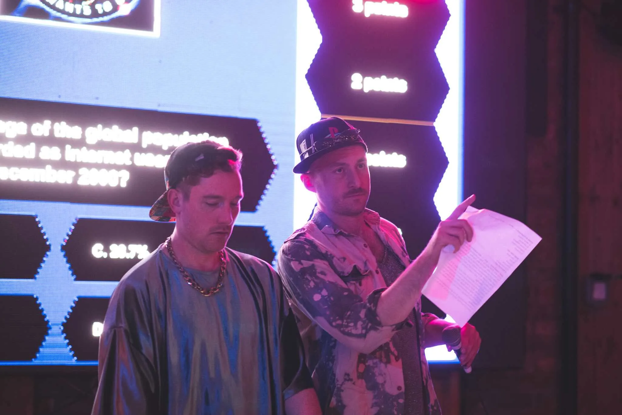 Two hosts presenting a quiz question with the scores board behind.